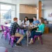 Four young adults sitting on purple chairs around a table studying together in a bright, modern lounge area.