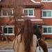 Two women with long hair stand outside a brick apartment building, one holding a smartphone and a white bag.