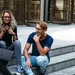 Three friends engaged in conversation on outdoor steps, sharing moments with a laptop and smartphone.