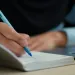 Close-up of hand writing notes in a notebook with a blue pen next to a laptop on a desk.