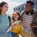 Group of diverse young adults smiling and walking outdoors on a sunny day with books and backpacks.