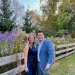 Smiling couple dressed formally standing on grass near wooden fence with colorful flowers in background