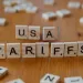 Wooden Scrabble tiles spelling USA TARIFFS on a wooden table with scattered tiles around.