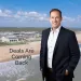 Smiling man in suit standing by beach resort with waves and text Deals Are Coming Back