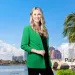 Smiling woman in green blazer stands by waterfront with palm trees and bridge in Palm Beach County, Florida.