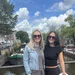 Two women in casual outfits and sunglasses standing by a canal with boats and trees in the background on a sunny day.