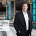 Man in blazer standing beside modern hotel bar with stools, glass lights, and backlit shelves.