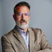 Confident middle-aged man with glasses and gray beard wearing a beige blazer and patterned shirt on a gray background