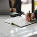 Person writing notes in a notebook at a white desk with a laptop and smartphone in the background.