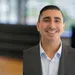 Smiling man in business suit with blurred office conference room and large windows in background.