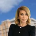 Confident businesswoman stands with folded arms in front of blurred city buildings under a blue sky.