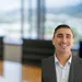 Smiling man in business suit with blurred office conference room and large windows in background.