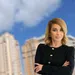 Confident businesswoman stands with folded arms in front of blurred city buildings under a blue sky.