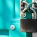 Close-up of a metal padlock securing a turquoise door with a metal latch in bright daylight.