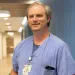 Male healthcare worker in blue scrubs writing on a clipboard in a hospital hallway with a phone in pocket.