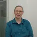 Woman with glasses and teal shirt speaking inside a modern office with glass walls and white background.