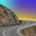 Winding mountain road with guardrails curving around rocky cliffs under a gradient sky.