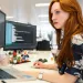 Focused woman with red hair working on coding displayed on dual monitors in a modern office environment.