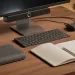 Modern workspace with monitor, wireless keyboard, notebook, pen, earbuds, and tech accessories on wooden desk.