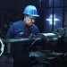 Worker in blue hard hat operating lathe machine in dimly lit industrial workshop focusing on metal cylinder