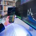Person holding smartphone calculator app with multiple monitors displaying financial charts and data