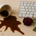 Spilled coffee next to a computer keyboard and a wilted red rose on a wooden desk surface.