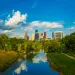 River reflecting clouds and city skyline with tall buildings and trees in vibrant daylight under blue sky.