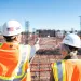 Two construction workers in hard hats and orange safety vests inspect rebar framework on a building site under clear sky.