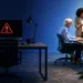 Office scene with mid-age and young professionals working, a computer displays a red warning triangle on a dark desk.