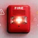 Red fire alarm bell with glowing light and warning triangles on textured wall background.