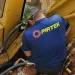 Worker in blue PIRTEK shirt repairs hydraulic machinery on yellow heavy equipment with various hoses and metal parts.