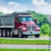 Red dump truck driving on rural road with green trees and blue sky background