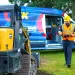 Construction worker in safety gear walking on grass near equipment and a service van with open side door.