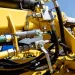 Close-up of yellow heavy machinery hydraulic system with metal pipes, valves, and hoses under bright daylight.