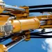 Close-up of yellow hydraulic machinery cylinders and pistons against a clear blue sky with clouds.