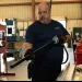 Man in safety glasses and gloves using a power tool on a metal pipe inside an industrial workshop.