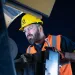 Worker wearing safety gear inspects machinery using a wrench under dim lighting conditions.