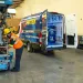 Technician in safety gear working on lift equipment next to a service van inside a spacious workshop.