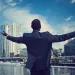 Businessman in suit stands on rooftop with arms outstretched overlooking city skyline under blue sky.