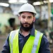 Confident male engineer in hard hat and reflective vest standing inside a factory or industrial setting.