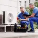 a couple of men sitting on a bench with a laptop and a fan
