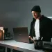 Man wearing beanie and black jacket doing cybercrime on a laptop at a metal desk under a desk lamp.