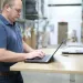 Man in blue shirt working on a laptop at a wooden table in a manufacturing warehouse with shelves and pallets.