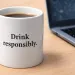 White coffee mug with Drink responsibly text next to a laptop on a wooden desk in a work setting