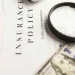 Magnifying glass on a cyber insurance policy document with US hundred-dollar bills partially visible beneath it.