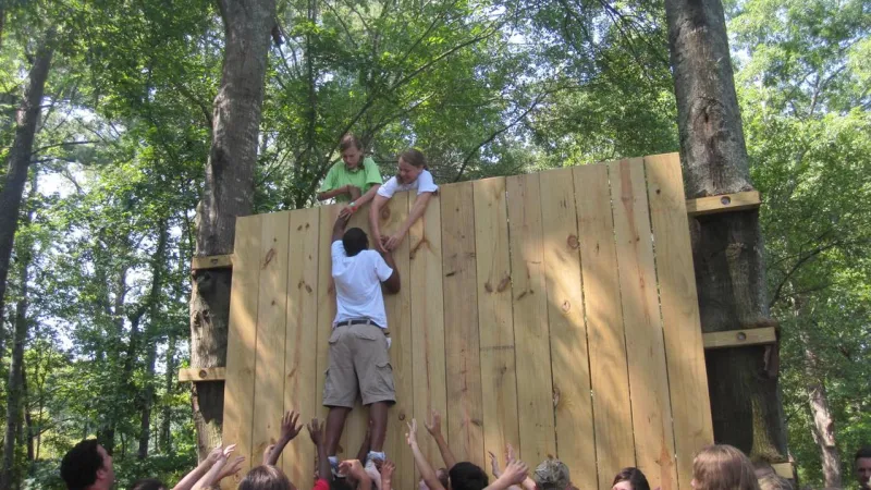 Group of people helping each other climb a tall wooden wall during an outdoor team-building activity.