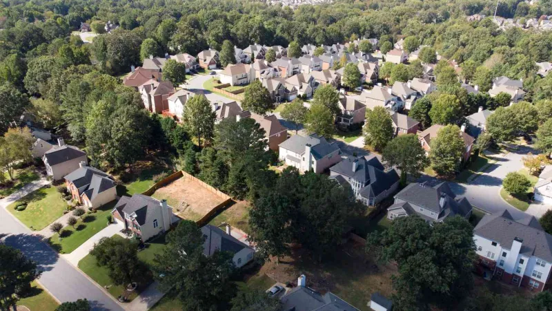 Aerial view of a suburban neighborhood with houses, trees, and roads surrounded by forested areas on a clear day