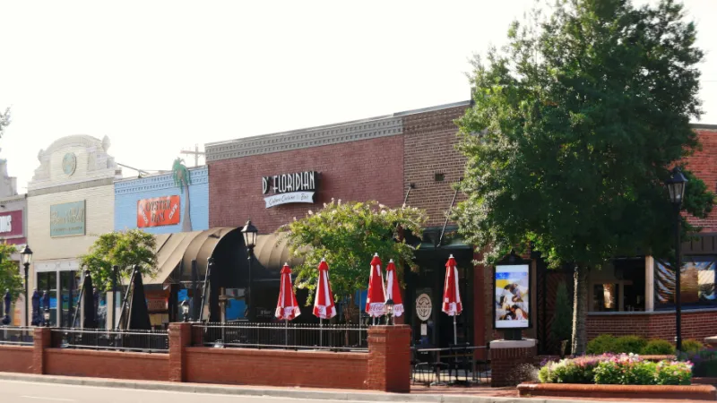 Street view of a brick restaurant with outdoor patio seating and red umbrellas on a sunny day.