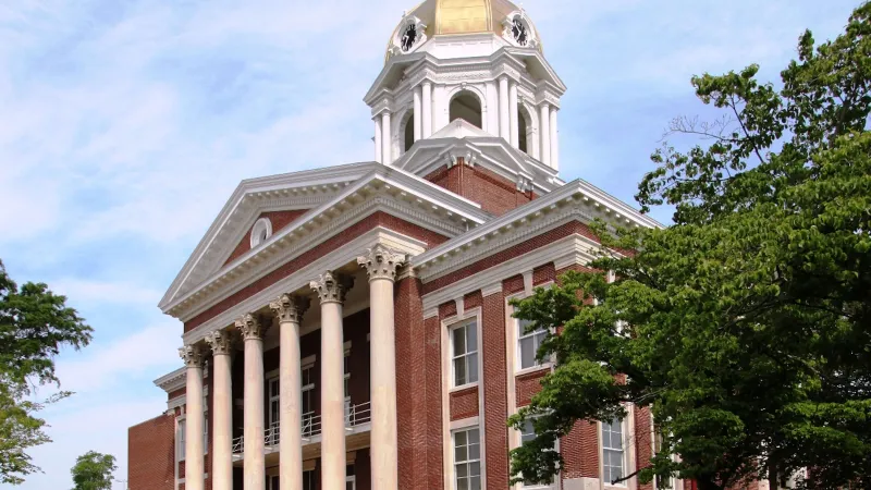 Historic red brick courthouse with tall white columns and golden dome under blue sky with trees