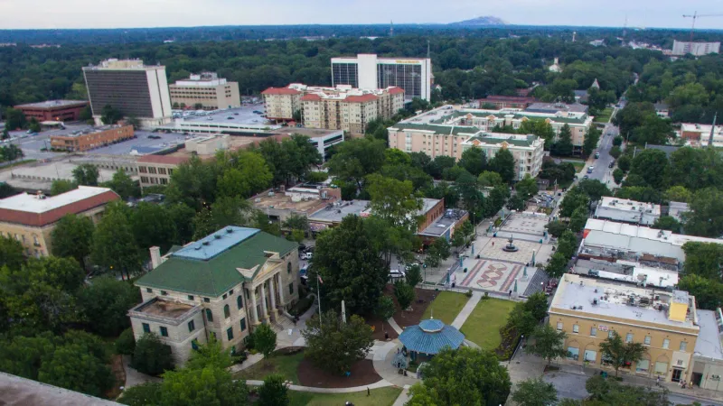 Aerial view of a city park with historic buildings, green trees, and surrounding urban structures under clear skies